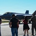 A group of observers wearing hearing protection stands on a flight line watching an F-35 Lightning II fighter jet taxi on the runway. The aircraft displays 'TX' tail codes indicating it is assigned to a Texas-based unit, with a partial tail number '5787' visible. One person in a red outfit appears to be waving at the aircraft while others observe.