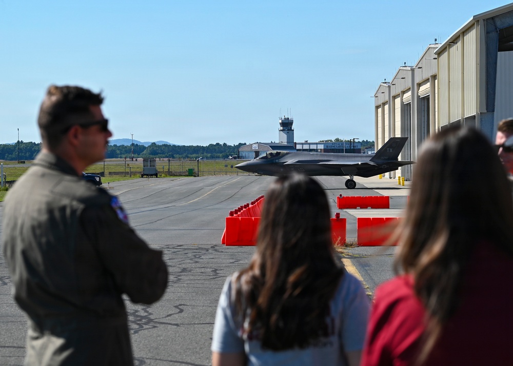 A military pilot in a flight suit and two civilians observe an F-35 stealth fighter jet taxiing on the flight line at what appears to be Barnes Regional Airport or a similar Air National Guard base. The scene is set on a clear, sunny day with hangars, orange safety barriers, and an air traffic control tower visible in the background. The perspective is from behind the observers, creating a depth-of-field composition with the aircraft in the midground.