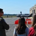 A military pilot in a flight suit and two civilians observe an F-35 stealth fighter jet taxiing on the flight line at what appears to be Barnes Regional Airport or a similar Air National Guard base. The scene is set on a clear, sunny day with hangars, orange safety barriers, and an air traffic control tower visible in the background. The perspective is from behind the observers, creating a depth-of-field composition with the aircraft in the midground.
