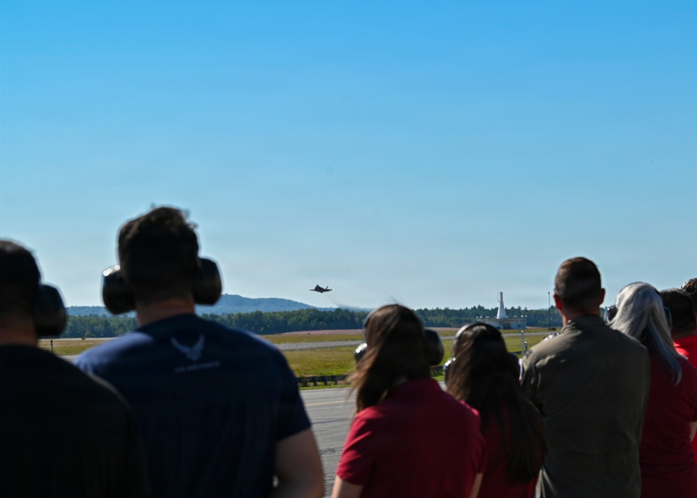 A group of spectators wearing hearing protection are watching a military jet aircraft performing a low flyover at what appears to be an airshow or military base open house. The observers are positioned on a tarmac or viewing area, facing the runway where the aircraft is visible in the distance. A tall structure, possibly a control tower or monument, is visible on the right side of the frame.