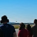 A group of spectators wearing hearing protection are watching a military jet aircraft performing a low flyover at what appears to be an airshow or military base open house. The observers are positioned on a tarmac or viewing area, facing the runway where the aircraft is visible in the distance. A tall structure, possibly a control tower or monument, is visible on the right side of the frame.