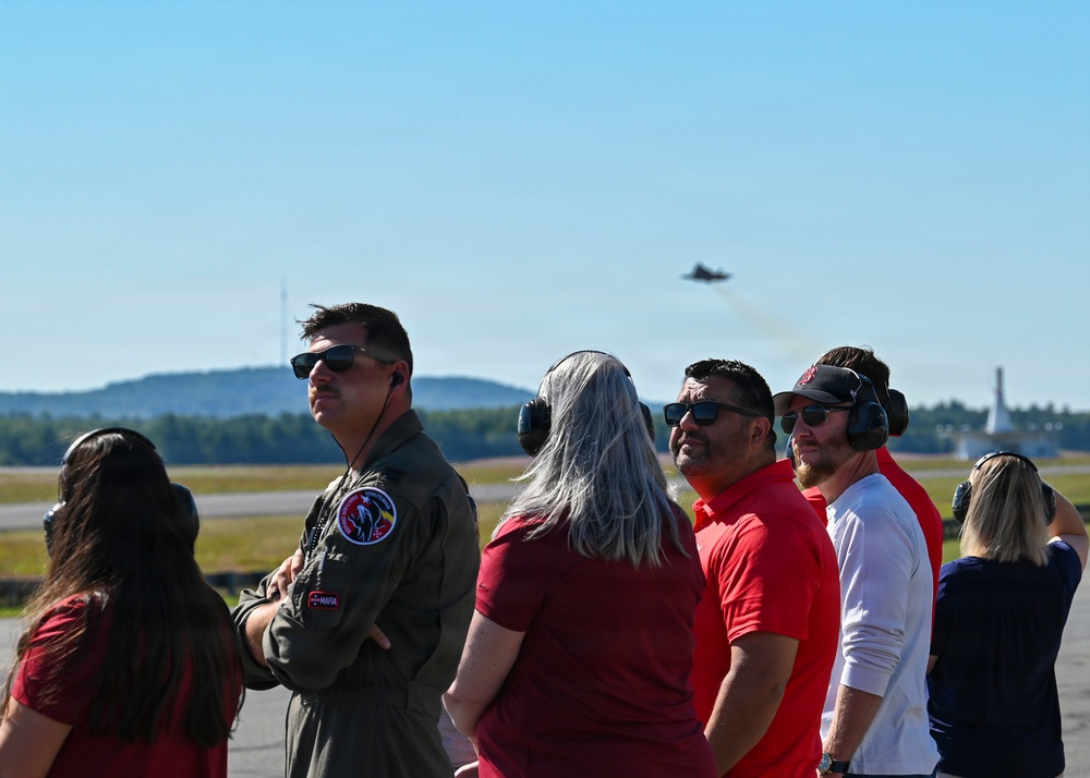 A group of civilians and at least one military pilot in a flight suit are observing aircraft operations on an airfield tarmac. All individuals are wearing hearing protection ear muffs, suggesting proximity to loud jet activity. At least one military jet aircraft is visible in the background sky, likely performing a demonstration or training flight.
