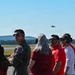 A group of civilians and at least one military pilot in a flight suit are observing aircraft operations on an airfield tarmac. All individuals are wearing hearing protection ear muffs, suggesting proximity to loud jet activity. At least one military jet aircraft is visible in the background sky, likely performing a demonstration or training flight.