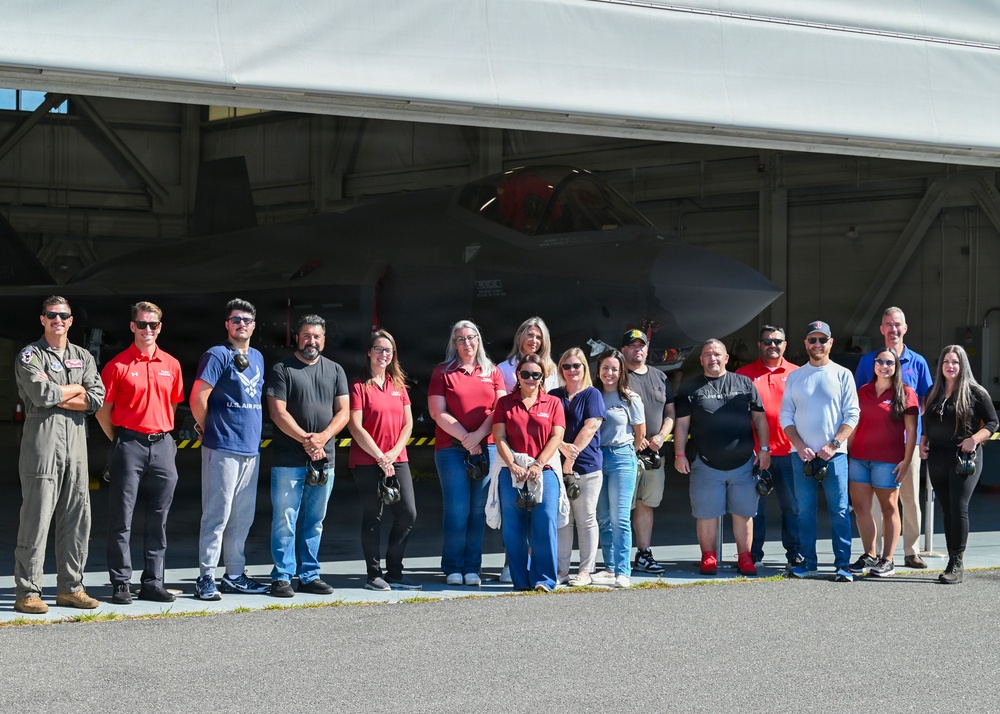A group of approximately 17 civilians and one uniformed U.S. Air Force pilot pose for a group photo in front of a military aircraft hangar. The aircraft visible in the background appears to be an F-35 or similar modern jet fighter partially housed in the hangar. This appears to be an official base tour or community outreach event.