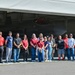 A group of approximately 17 civilians and one uniformed U.S. Air Force pilot pose for a group photo in front of a military aircraft hangar. The aircraft visible in the background appears to be an F-35 or similar modern jet fighter partially housed in the hangar. This appears to be an official base tour or community outreach event.