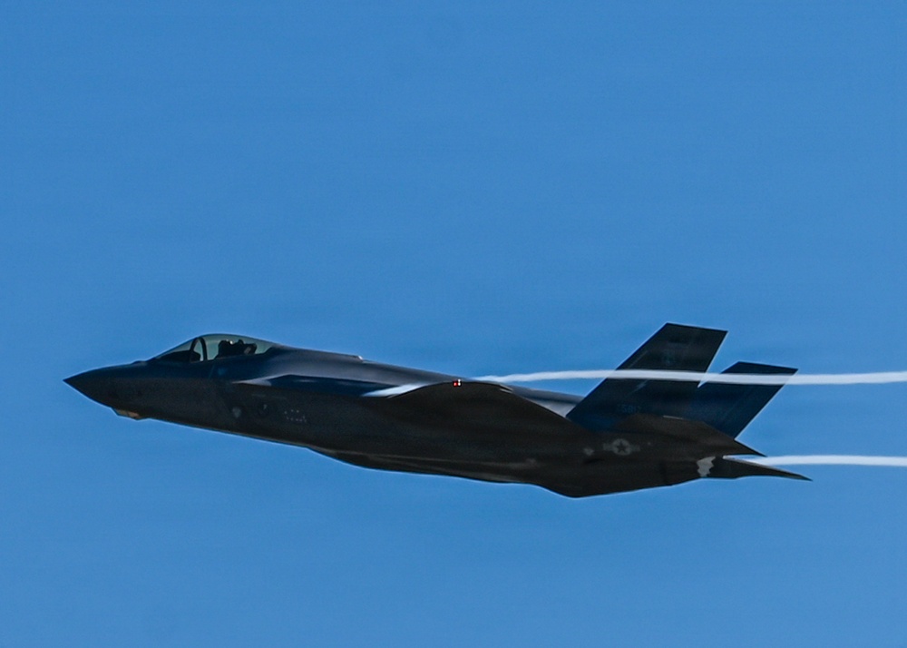 A dark-colored fifth-generation stealth fighter jet, consistent with an F-35 Lightning II, is captured in level flight against a clear blue sky. The aircraft is producing visible wingtip or wing-root condensation trails (contrails). The image is sharp and well-exposed, providing excellent detail of the aircraft's stealth airframe geometry.