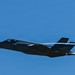 A dark-colored fifth-generation stealth fighter jet, consistent with an F-35 Lightning II, is captured in level flight against a clear blue sky. The aircraft is producing visible wingtip or wing-root condensation trails (contrails). The image is sharp and well-exposed, providing excellent detail of the aircraft's stealth airframe geometry.