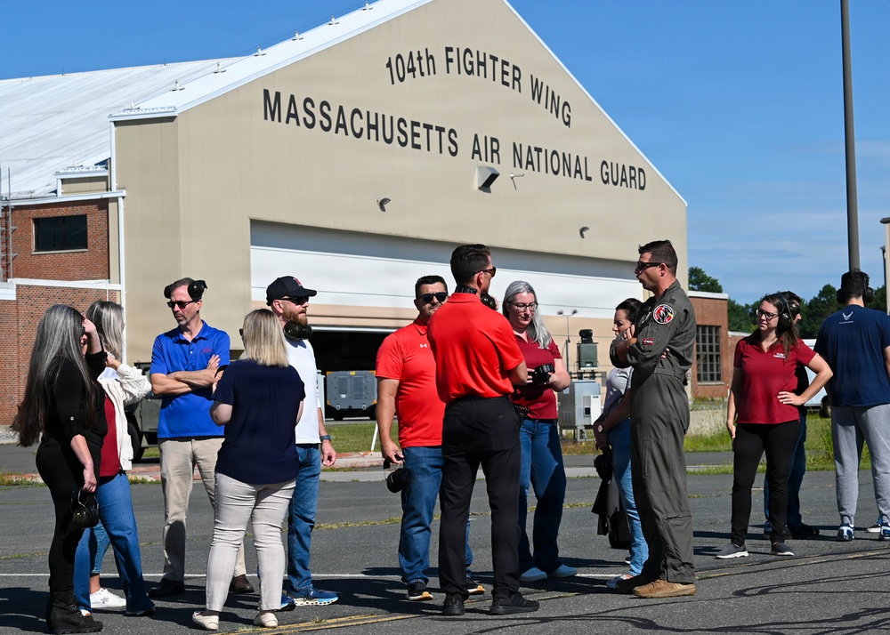 A group of approximately 12 civilians and one military pilot in a flight suit are gathered outside a large hangar at Barnes Air National Guard Base in Massachusetts. The hangar prominently displays signage reading '104th Fighter Wing, Massachusetts Air National Guard.' The gathering appears to be an organized visit or briefing, possibly a community outreach or media event.