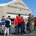 A group of approximately 12 civilians and one military pilot in a flight suit are gathered outside a large hangar at Barnes Air National Guard Base in Massachusetts. The hangar prominently displays signage reading '104th Fighter Wing, Massachusetts Air National Guard.' The gathering appears to be an organized visit or briefing, possibly a community outreach or media event.