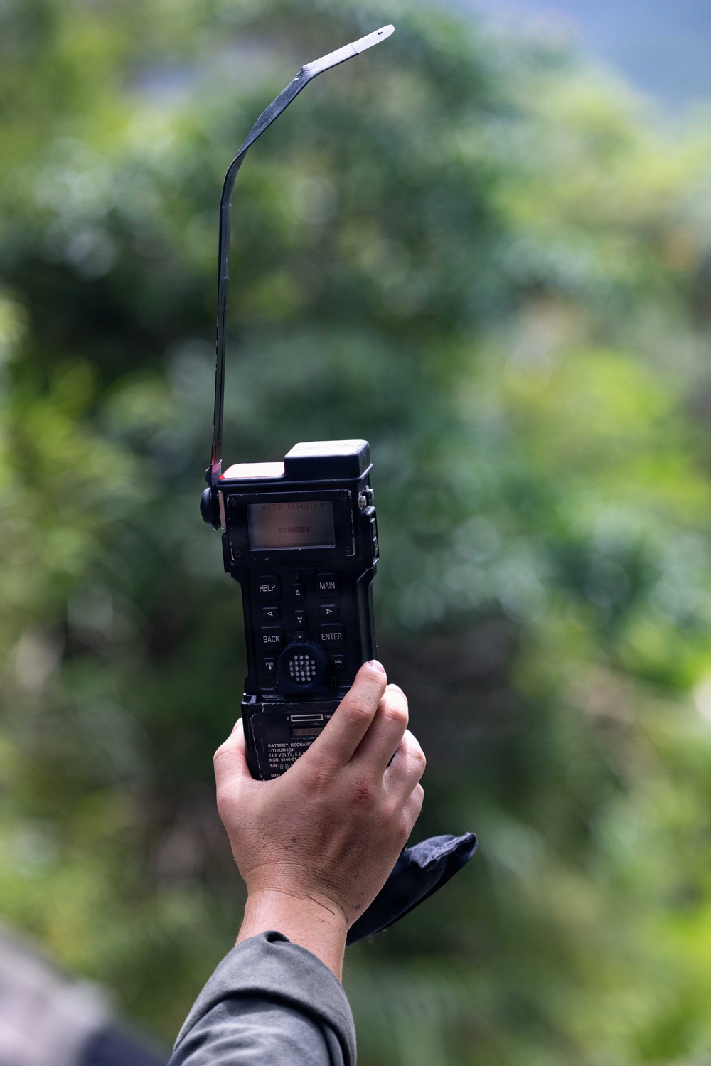 A hand is holding up a black military-style handheld radio or communication device with a long, bent whip antenna extended upward. The device displays 'STANDBY' on its small LCD screen and features labeled buttons including HELP, MAIN, BACK, and ENTER. The background consists of blurred green foliage and trees, suggesting an outdoor environment.
