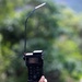 A hand is holding up a black military-style handheld radio or communication device with a long, bent whip antenna extended upward. The device displays 'STANDBY' on its small LCD screen and features labeled buttons including HELP, MAIN, BACK, and ENTER. The background consists of blurred green foliage and trees, suggesting an outdoor environment.