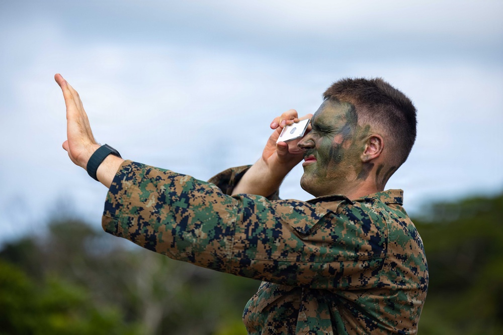A military personnel wearing MARPAT (Marine pattern) digital camouflage uniform and tactical face paint is talking on a smartphone while raising one hand in a 'stop' or signaling gesture. The individual appears to be outdoors in a field or training environment with vegetation and overcast sky visible in the background. The combination of combat face paint and mobile phone use suggests a field exercise or training scenario.