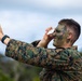 A military personnel wearing MARPAT (Marine pattern) digital camouflage uniform and tactical face paint is talking on a smartphone while raising one hand in a 'stop' or signaling gesture. The individual appears to be outdoors in a field or training environment with vegetation and overcast sky visible in the background. The combination of combat face paint and mobile phone use suggests a field exercise or training scenario.