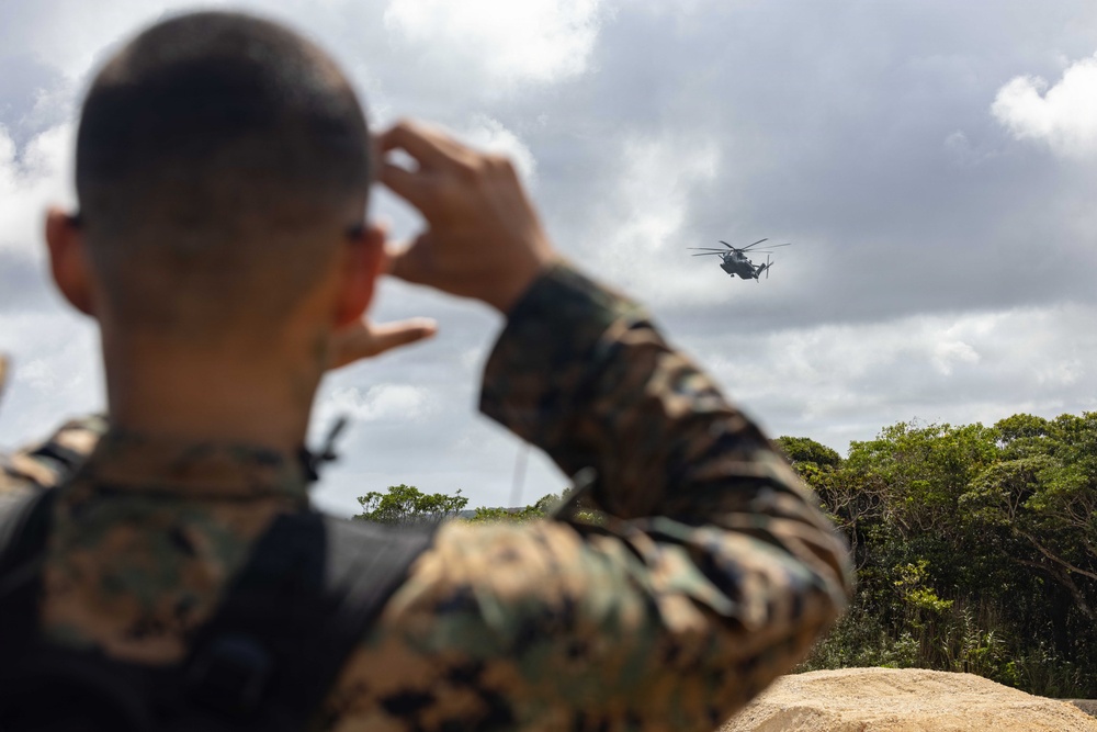 A military service member in camouflage uniform is observed from behind, shielding their eyes while watching a large helicopter flying in the distance. The scene appears to take place in a tropical or subtropical outdoor environment with dense green vegetation. The image captures what appears to be a military training or operational exercise involving rotary-wing aircraft.