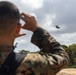 A military service member in camouflage uniform is observed from behind, shielding their eyes while watching a large helicopter flying in the distance. The scene appears to take place in a tropical or subtropical outdoor environment with dense green vegetation. The image captures what appears to be a military training or operational exercise involving rotary-wing aircraft.
