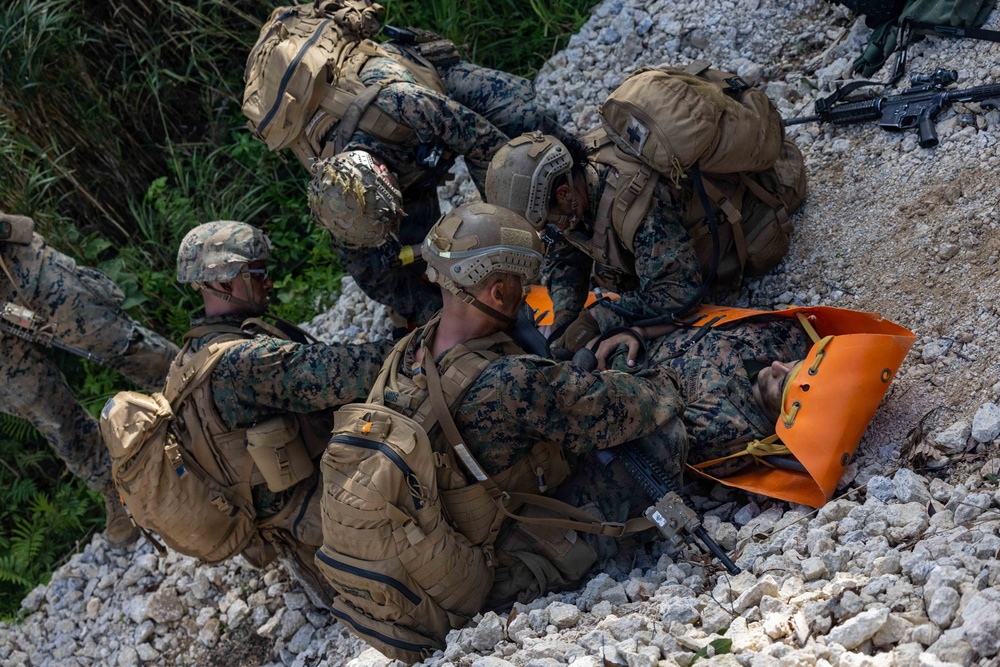 Multiple military personnel in MARPAT (Marine pattern) camouflage uniforms are conducting a casualty evacuation (CASEVAC) drill on a rocky terrain. Several Marines are attending to a simulated casualty lying on an orange litter/stretcher. The scene appears to be a training exercise based on the controlled environment and use of a brightly colored rescue litter.