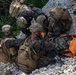 Multiple military personnel in MARPAT (Marine pattern) camouflage uniforms are conducting a casualty evacuation (CASEVAC) drill on a rocky terrain. Several Marines are attending to a simulated casualty lying on an orange litter/stretcher. The scene appears to be a training exercise based on the controlled environment and use of a brightly colored rescue litter.