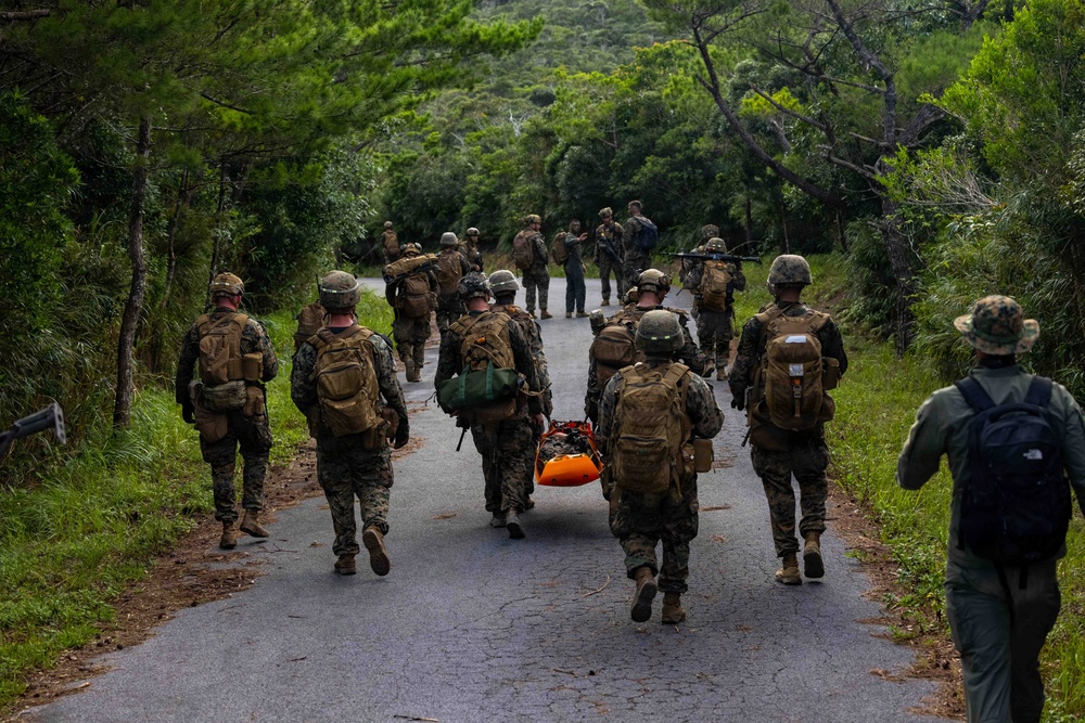 A group of military personnel in camouflage uniforms and tactical gear are moving along a narrow paved road through a lush, green forested area. Two individuals in the center are carrying an orange rescue stretcher or litter. A civilian or observer in civilian clothing with a blue backpack walks alongside the group on the right.