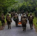 A group of military personnel in camouflage uniforms and tactical gear are moving along a narrow paved road through a lush, green forested area. Two individuals in the center are carrying an orange rescue stretcher or litter. A civilian or observer in civilian clothing with a blue backpack walks alongside the group on the right.