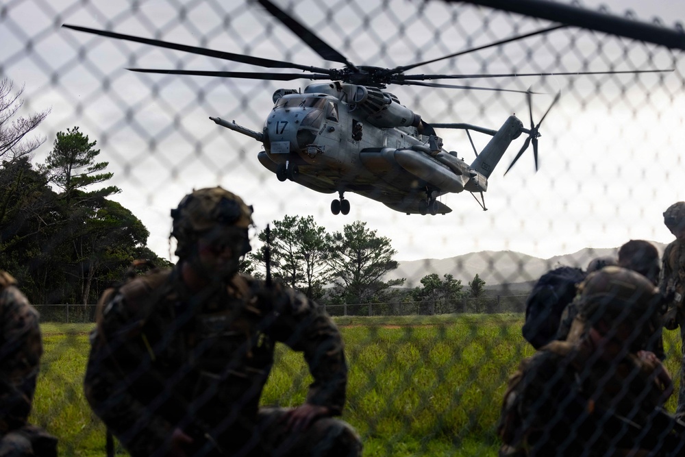 Military personnel in camouflage uniforms and tactical gear observe a large military helicopter numbered '17' hovering low over a grassy field. The scene appears to be a training exercise or operational insertion, with soldiers positioned behind a chain-link fence watching the aircraft. The helicopter is consistent with a Sikorsky CH-53E Super Stallion used by the U.S. Marine Corps.