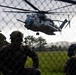 Military personnel in camouflage uniforms and tactical gear observe a large military helicopter numbered '17' hovering low over a grassy field. The scene appears to be a training exercise or operational insertion, with soldiers positioned behind a chain-link fence watching the aircraft. The helicopter is consistent with a Sikorsky CH-53E Super Stallion used by the U.S. Marine Corps.