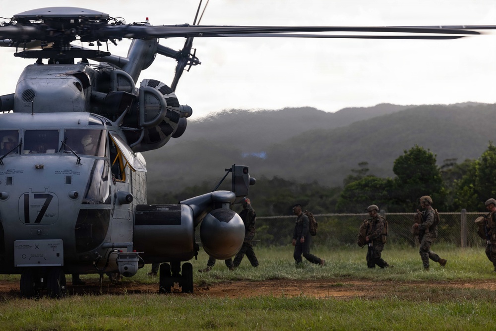 A large military helicopter, marked with the number '17', is parked on a grassy field while several soldiers in combat gear approach or board the aircraft. The setting appears to be a tropical or subtropical environment, with dense green hills visible in the background under overcast skies. This appears to be a military exercise or operational deployment involving rotary-wing aviation assets.