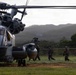 A large military helicopter, marked with the number '17', is parked on a grassy field while several soldiers in combat gear approach or board the aircraft. The setting appears to be a tropical or subtropical environment, with dense green hills visible in the background under overcast skies. This appears to be a military exercise or operational deployment involving rotary-wing aviation assets.