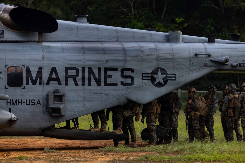 A group of U.S. Marines in combat gear are gathered alongside a large military helicopter marked 'MARINES' and 'HMH-USA'. The aircraft appears to be a CH-53 heavy-lift helicopter, identifiable by its tandem rotor configuration and fuselage shape. Personnel appear to be preparing for or completing a helicopter insertion/extraction operation in a field environment.