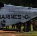 A group of U.S. Marines in combat gear are gathered alongside a large military helicopter marked 'MARINES' and 'HMH-USA'. The aircraft appears to be a CH-53 heavy-lift helicopter, identifiable by its tandem rotor configuration and fuselage shape. Personnel appear to be preparing for or completing a helicopter insertion/extraction operation in a field environment.