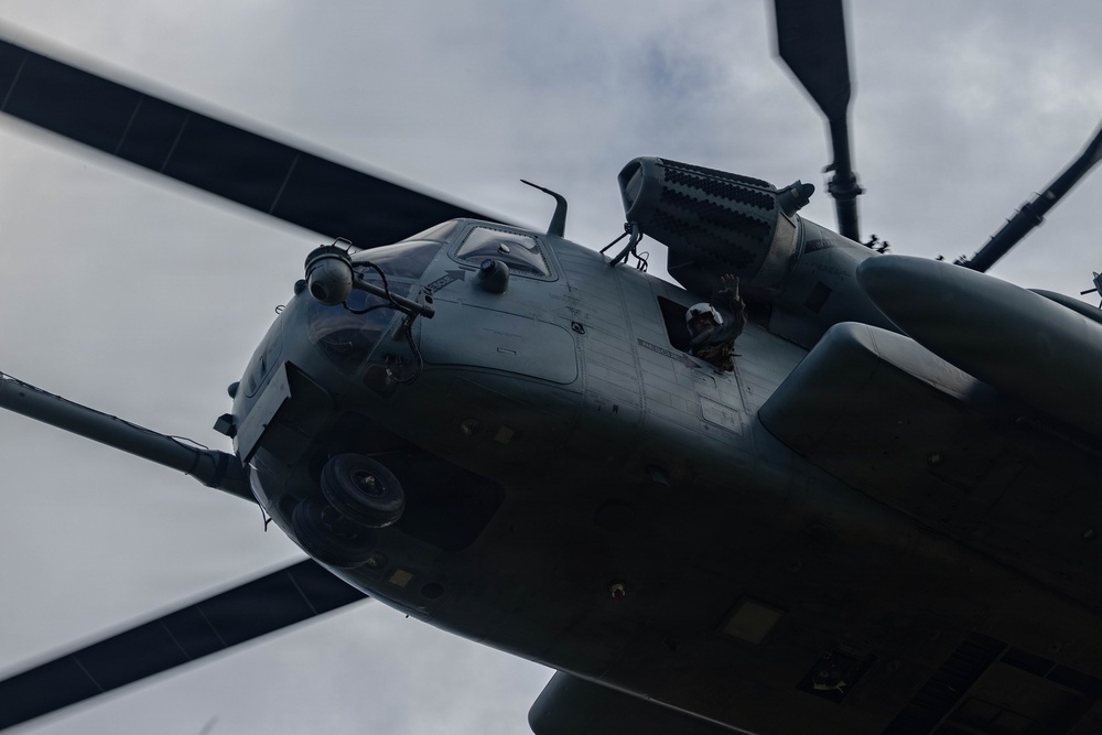 A low-angle upward shot of a large military helicopter, likely a Sikorsky CH-53 or similar heavy-lift variant, photographed from directly below. The image captures the nose section, cockpit glazing, rotor blades, and engine housing in close detail. A crew member is visible leaning out of an open window or door on the right side of the fuselage.
