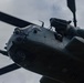 A low-angle upward shot of a large military helicopter, likely a Sikorsky CH-53 or similar heavy-lift variant, photographed from directly below. The image captures the nose section, cockpit glazing, rotor blades, and engine housing in close detail. A crew member is visible leaning out of an open window or door on the right side of the fuselage.