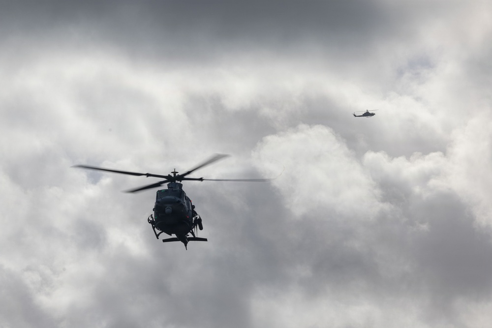 Two military helicopters are flying in formation against a heavily overcast, dramatic sky. The foreground helicopter is a dark-colored, small scout or light attack helicopter with visible rotor blur indicating active flight. A second helicopter is visible in the upper right background at a greater distance.