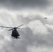 Two military helicopters are flying in formation against a heavily overcast, dramatic sky. The foreground helicopter is a dark-colored, small scout or light attack helicopter with visible rotor blur indicating active flight. A second helicopter is visible in the upper right background at a greater distance.