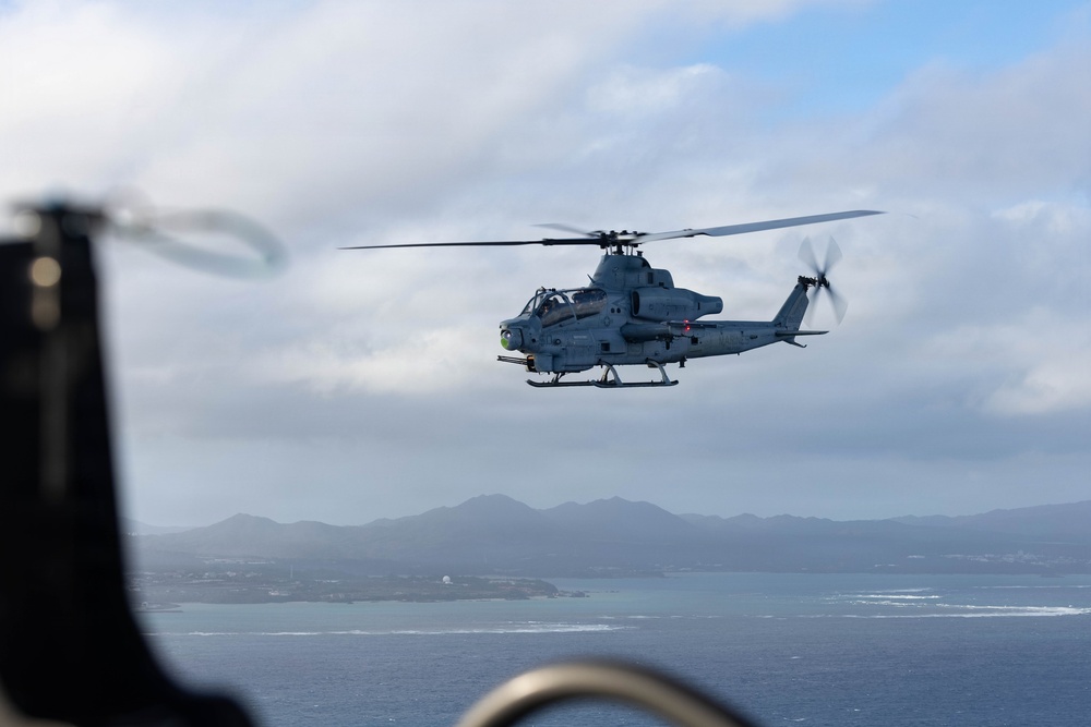 A military attack helicopter, identifiable as a Bell AH-1Z Viper, is photographed in flight over a coastal area with islands and mountains visible in the background. The image appears to be taken from another aircraft, with blurred foreground elements suggesting the photographer is aboard a nearby helicopter or aircraft. The gray paint scheme and weapons systems indicate this is a U.S. Marine Corps combat helicopter.