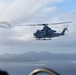 A military attack helicopter, identifiable as a Bell AH-1Z Viper, is photographed in flight over a coastal area with islands and mountains visible in the background. The image appears to be taken from another aircraft, with blurred foreground elements suggesting the photographer is aboard a nearby helicopter or aircraft. The gray paint scheme and weapons systems indicate this is a U.S. Marine Corps combat helicopter.