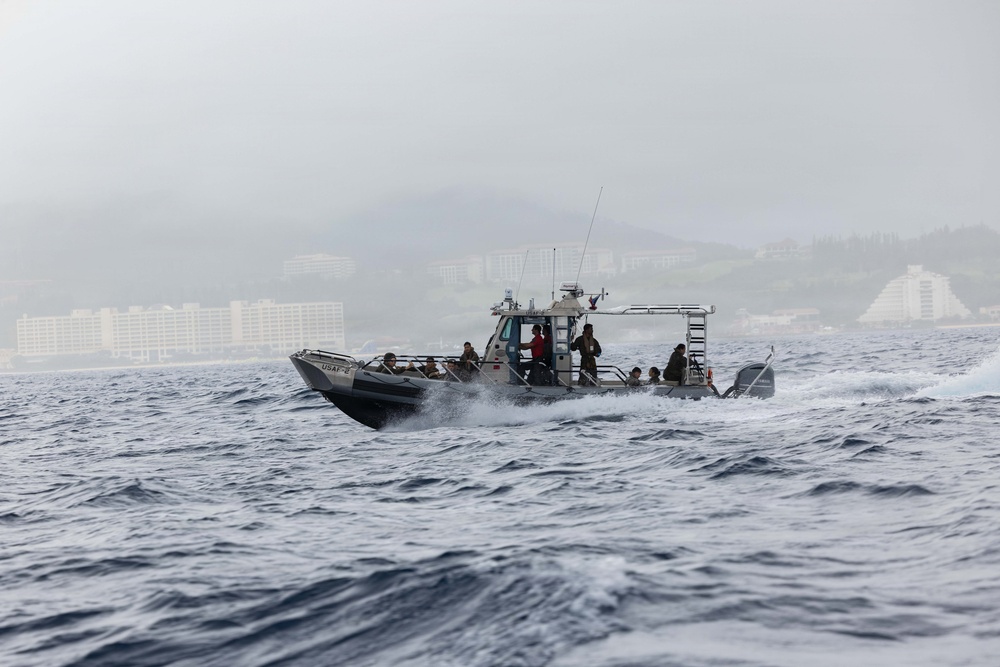 A military patrol boat marked 'USAF' is underway at speed on choppy open water, carrying several personnel in military-style attire. The vessel appears to be a rigid-hull inflatable boat (RHIB) or similar tactical craft operating near a coastal area with visible buildings and hills in the background. Weather conditions appear overcast and hazy.