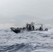 A military patrol boat marked 'USAF' is underway at speed on choppy open water, carrying several personnel in military-style attire. The vessel appears to be a rigid-hull inflatable boat (RHIB) or similar tactical craft operating near a coastal area with visible buildings and hills in the background. Weather conditions appear overcast and hazy.