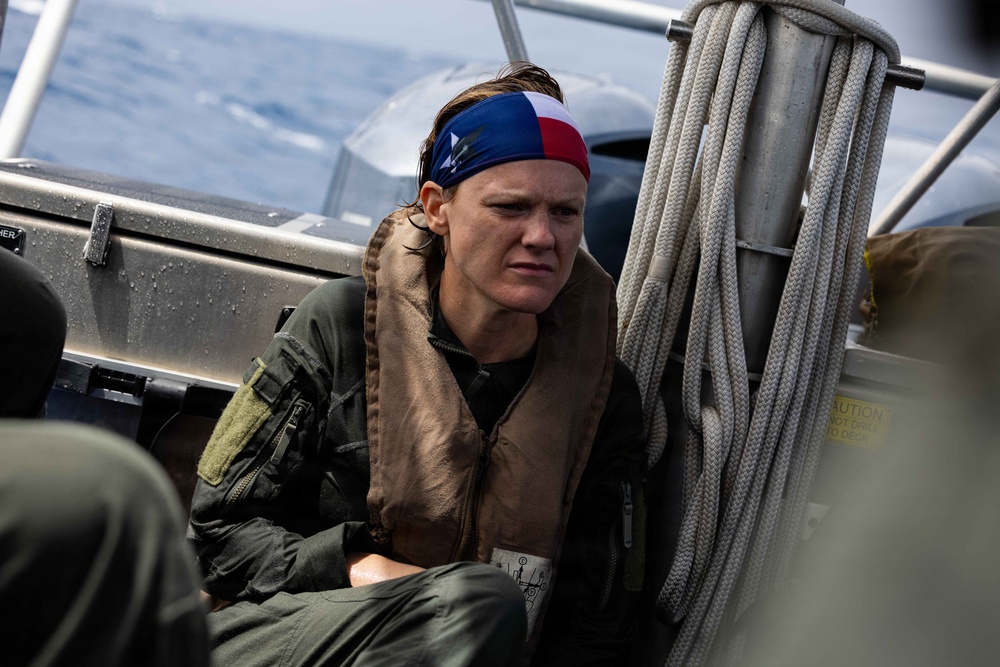 A person wearing a military flight suit and life vest sits aboard a watercraft at sea. They are wearing a red, white, and blue bandana/headband and appear wet, suggesting recent water immersion or exposure. The setting and attire suggest a military maritime rescue or survival training exercise.