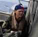 A person wearing a military flight suit and life vest sits aboard a watercraft at sea. They are wearing a red, white, and blue bandana/headband and appear wet, suggesting recent water immersion or exposure. The setting and attire suggest a military maritime rescue or survival training exercise.