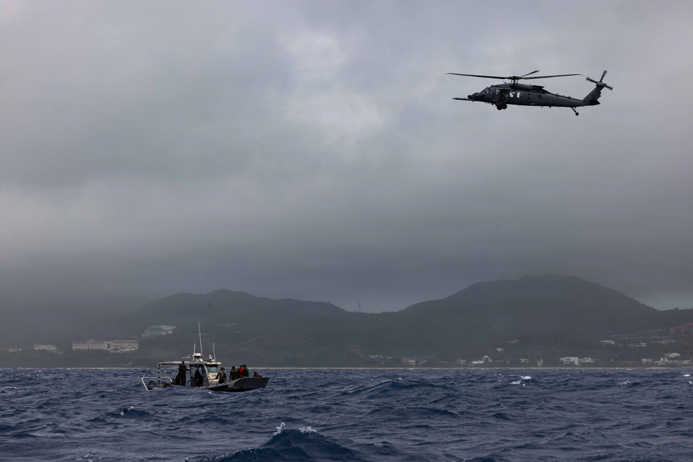 A military or rescue helicopter flies over choppy ocean waters near a coastline with mountainous terrain in the background. Two small vessels, likely patrol or rescue boats, are visible on the water below the helicopter. The scene appears to take place under overcast, stormy weather conditions.