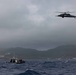 A military or rescue helicopter flies over choppy ocean waters near a coastline with mountainous terrain in the background. Two small vessels, likely patrol or rescue boats, are visible on the water below the helicopter. The scene appears to take place under overcast, stormy weather conditions.