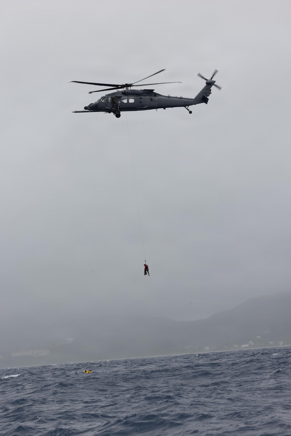 A military helicopter, consistent with an HH-60 Pave Hawk or MH-60 Seahawk variant, is hovering over open ocean water while conducting a hoist operation. A person in a red garment is suspended on a cable below the aircraft, positioned above the water surface. A yellow flotation device or marker is visible on the water surface below.
