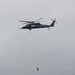 A military helicopter, consistent with an HH-60 Pave Hawk or MH-60 Seahawk variant, is hovering over open ocean water while conducting a hoist operation. A person in a red garment is suspended on a cable below the aircraft, positioned above the water surface. A yellow flotation device or marker is visible on the water surface below.
