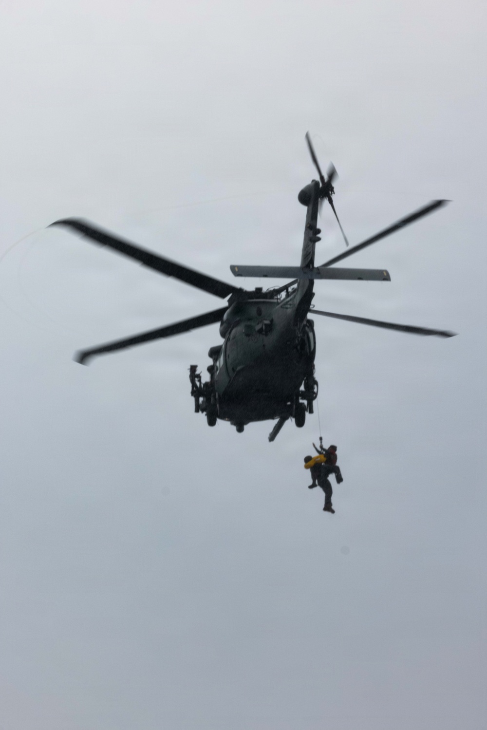 A dark military or rescue helicopter is hovering in an overcast sky while conducting a hoist operation. One or two personnel are suspended below the aircraft on a cable, appearing to be in the process of being lifted or lowered. The image captures an active search-and-rescue or training operation.