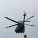A dark military or rescue helicopter is hovering in an overcast sky while conducting a hoist operation. One or two personnel are suspended below the aircraft on a cable, appearing to be in the process of being lifted or lowered. The image captures an active search-and-rescue or training operation.