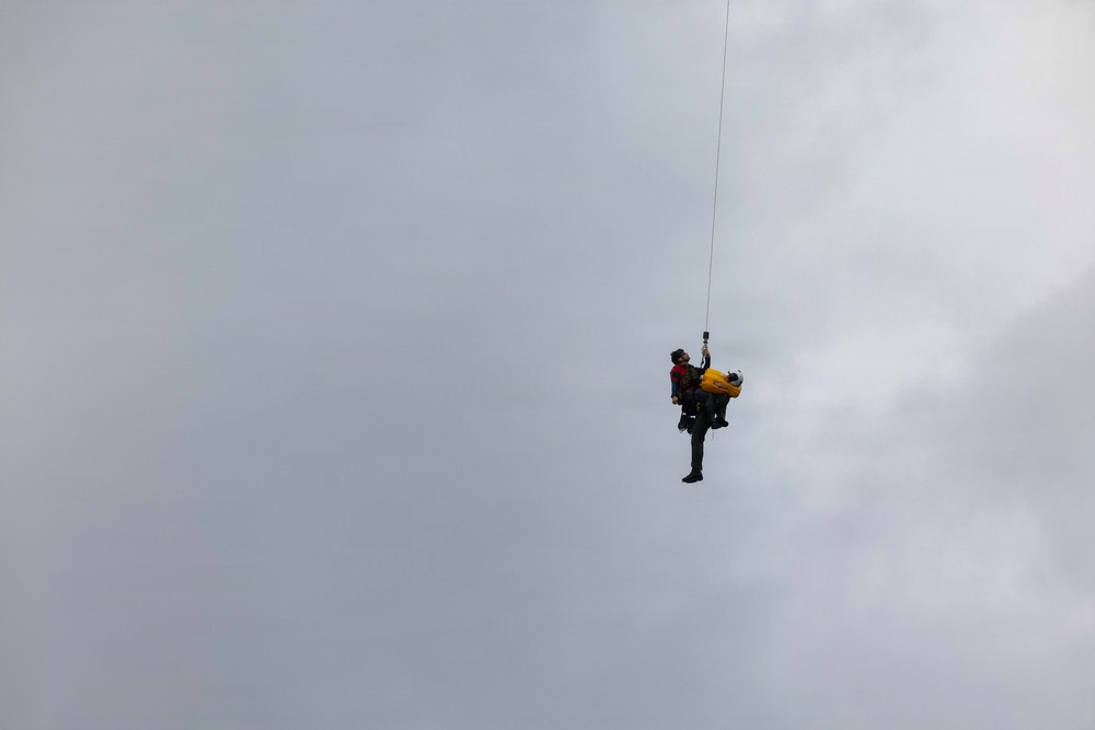 Two individuals are suspended mid-air from a single rope or cable extending upward out of frame, suggesting a helicopter hoist or rescue operation. One person appears to be a rescuer in dark clothing with a red vest, while the other appears to be a casualty or passenger wearing a yellow pack or harness. The scene takes place against an overcast, grey sky.