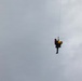 Two individuals are suspended mid-air from a single rope or cable extending upward out of frame, suggesting a helicopter hoist or rescue operation. One person appears to be a rescuer in dark clothing with a red vest, while the other appears to be a casualty or passenger wearing a yellow pack or harness. The scene takes place against an overcast, grey sky.