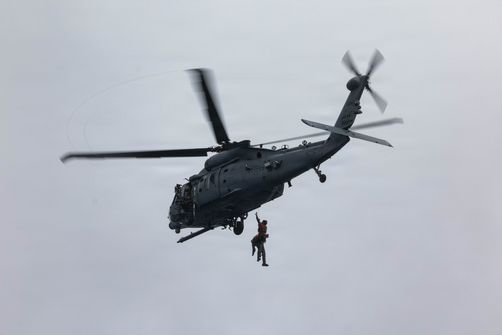 A military helicopter, appearing to be an HH-60 Pave Hawk or similar variant, is hovering in an overcast sky while conducting a hoist or rescue operation. A person in an orange survival suit or rescue gear is suspended below the aircraft on a cable or hoist line. The helicopter displays U.S. military markings and is painted in a dark gray tactical color scheme.