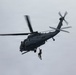 A military helicopter, appearing to be an HH-60 Pave Hawk or similar variant, is hovering in an overcast sky while conducting a hoist or rescue operation. A person in an orange survival suit or rescue gear is suspended below the aircraft on a cable or hoist line. The helicopter displays U.S. military markings and is painted in a dark gray tactical color scheme.