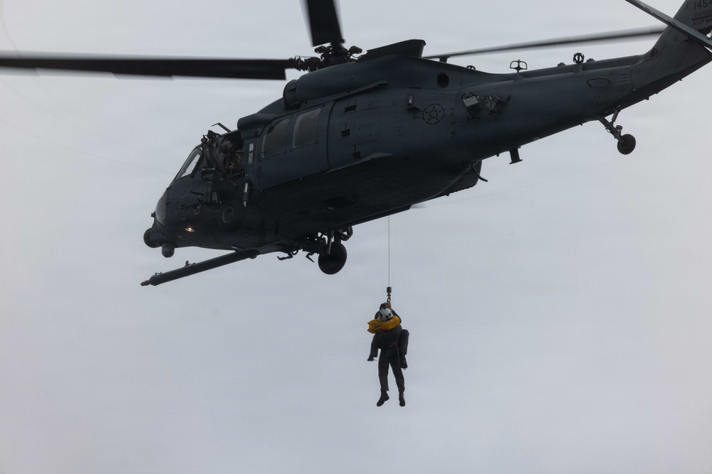 A dark gray military helicopter, identifiable as a U.S. Air Force HH-60 Pave Hawk, is hovering against an overcast sky. A person is being hoisted on a cable beneath the aircraft, wearing a yellow flotation device and helmet, indicating a search and rescue or combat rescue training operation. The scene depicts a hoist/rescue extraction exercise or live rescue mission.